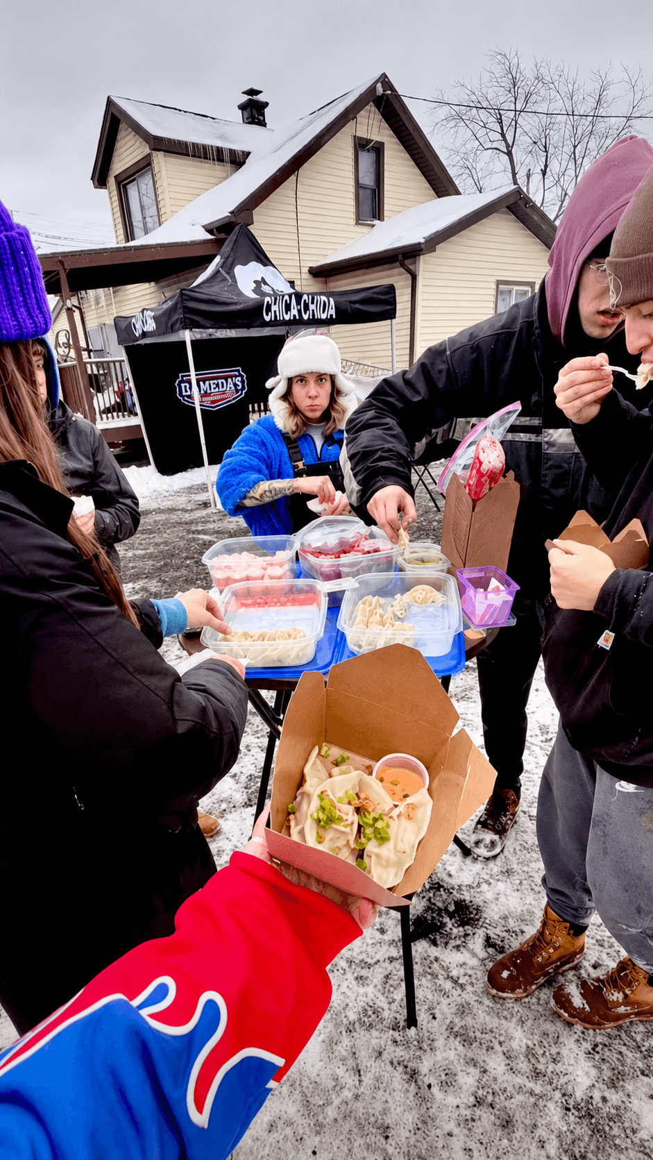 A fan in a Buffalo Bills jacket holding up a box of Daughters Dumplings at a snowy tailgate